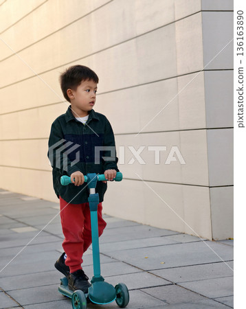 Young Child Riding A Scooter Down A Sleek, Modern Walkway. 136163890