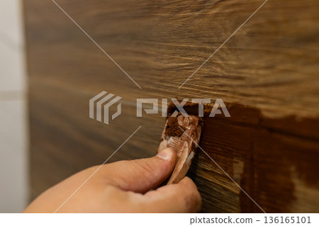 A person applies grout with a putty knife into the cracks between wood-look ceramic tiles during a DIY home renovation. A close-up of hands finishing the grout joints on a bathroom wall. A person applies grout with a putty knife into the cracks between wood-look ceramic tiles during a DIY home renovation. A close-up of hands finishing the grout joints on a bathroom wall. 136165101
