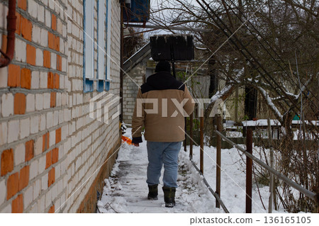 Man walks along a snowy path beside a brick house carrying a shovel on his shoulder, heading out to clear winter snow from the yard and perform daily outdoor chores. 136165105