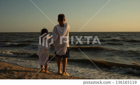 Two young siblings stand hand in hand on a sandy beach at sunset, gazing toward the calm sea and horizon, sharing a peaceful, golden-hour moment of togetherness and hope. Two young siblings stand hand in hand on a sandy beach at sunset, gazing toward the calm sea and horizon, sharing a peaceful, golden-hour moment of togetherness and hope. 136165119