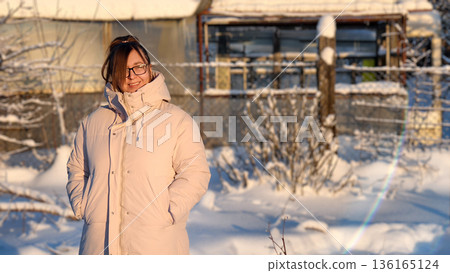 A young woman wearing glasses and a warm jacket stands in a snowy courtyard on a sunny winter day. She smiles calmly and enjoys the peaceful, frosty landscape and soft daylight. 136165124