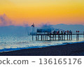 A Serene Sunset Over a Pier with Silhouetted Figures, Vibrant Colors Reflected in Water, and a Calm Beach Setting in the Background 136165176