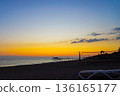 A Serene Beach Scene at Sunset with Vibrant Colors in the Sky Reflecting Over Calm Waters and Silhouetted Structures Along the Shoreline 136165177