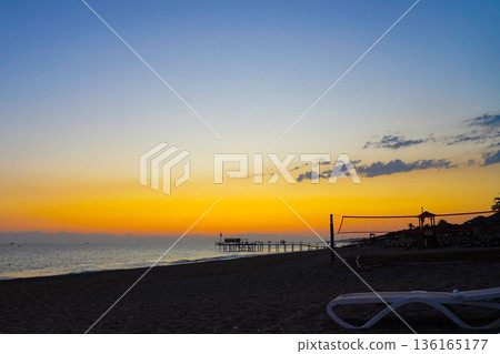 A Serene Beach Scene at Sunset with Vibrant Colors in the Sky Reflecting Over Calm Waters and Silhouetted Structures Along the Shoreline A Serene Beach Scene at Sunset with Vibrant Colors in the Sky Reflecting Over Calm Waters and Silhouetted Structures Along the Shoreline 136165177