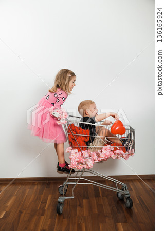 Kids play in shopping cart with flowers on the side in a simple room with wooden floor 136165924