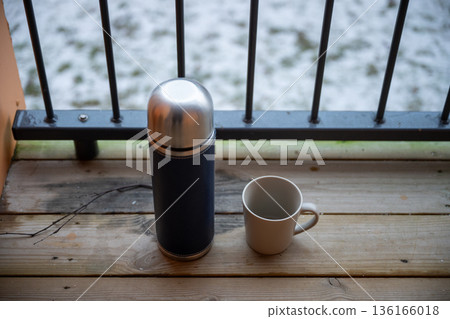 Thermos flask and ceramic cup on wooden balcony with snowy winter background Thermos flask and ceramic cup on wooden balcony with snowy winter background 136166018