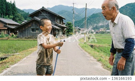 一個調皮的男孩在鄉間小路上用軟管向他的祖父噴水。 一個調皮的男孩在鄉間小路上用軟管向他的祖父噴水。 136166193