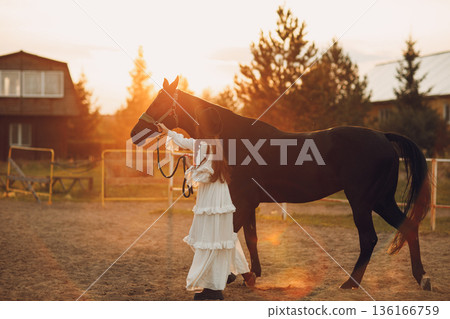 Woman in white dress and black hat with her horse at sunset outdoors ranch 136166759