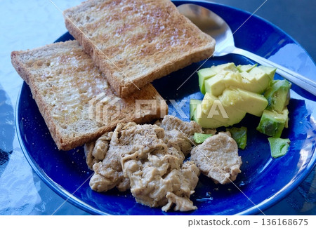 A nutritious morning meal featuring crispy whole wheat toast slices, fresh diced green avocado, and seasoned scrambled eggs served on a vibrant blue ceramic plate. 136168675