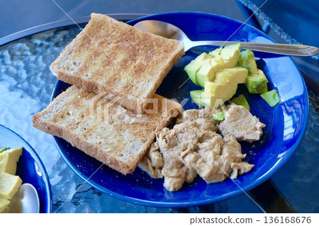 A vibrant blue plate holds a satisfying, healthy plant based meal featuring golden brown buttered toast, creamy sliced avocado, and a savory protein portion, perfect for breakfast or brunch. 136168676