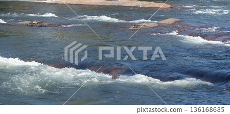 The beautiful blue river water flows dynamically over and around various rocks, creating picturesque white rapids and gentle ripples across the surface in a natural stream. The beautiful blue river water flows dynamically over and around various rocks, creating picturesque white rapids and gentle ripples across the surface in a natural stream. 136168685