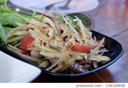 A vibrant close up of spicy Thai green papaya salad with shredded papaya fresh tomatoes chilies and cockles served in a black bowl with vegetables. 136168698