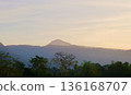 A tranquil mountain landscape featuring layers of misty forested hills silhouetted against a softly glowing sky during sunrise with vibrant green trees in the foreground. 136168707