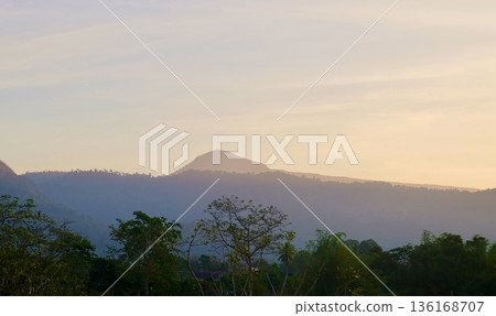 A tranquil mountain landscape featuring layers of misty forested hills silhouetted against a softly glowing sky during sunrise with vibrant green trees in the foreground. A tranquil mountain landscape featuring layers of misty forested hills silhouetted against a softly glowing sky during sunrise with vibrant green trees in the foreground. 136168707
