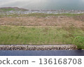 A high-angle shot captures distinct horizontal layers of a serene lakeside, featuring calm water, rocky shoreline, dry earth, lush green grass, and stone landscaping. 136168708