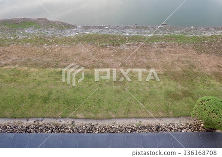 A high-angle shot captures distinct horizontal layers of a serene lakeside, featuring calm water, rocky shoreline, dry earth, lush green grass, and stone landscaping. A high-angle shot captures distinct horizontal layers of a serene lakeside, featuring calm water, rocky shoreline, dry earth, lush green grass, and stone landscaping. 136168708