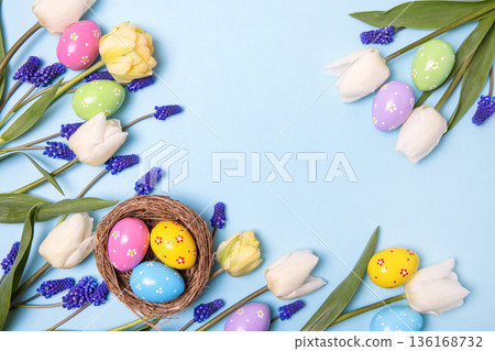 Easter colored painted eggs in bird nest with yellow daffodils, white tulips, and blue muscari on blue background with space for text and greetings. Easter and spring concept. Spring background Easter colored painted eggs in bird nest with yellow daffodils, white tulips, and blue muscari on blue background with space for text and greetings. Easter and spring concept. Spring background 136168732