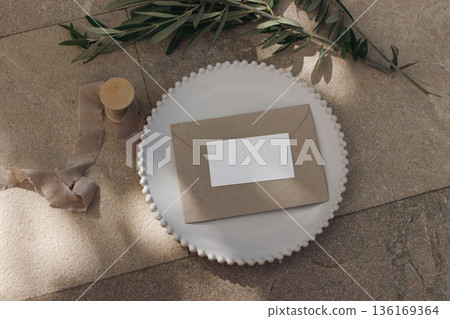 Wedding setting flat lay. Blank place business card, envelope on ceramic plate, olive tree branches, ribbon. Beige marble floor. Rustic Mediterranean summer dining scene. Stationery mockup copy space. 136169364