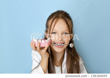 Smiling Girl holding dental model and mirror tool, symbolizing oral hygiene education, pediatric dentistry and World Oral Health Day awareness on blue background. 136169445