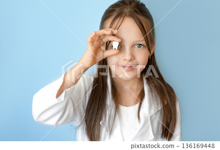 Smiling child holding a lost milk tooth near eye on blue background, symbol of first tooth loss, oral hygiene education, pediatric dentistry and World Oral Health Day awareness on March 20. Smiling child holding a lost milk tooth near eye on blue background, symbol of first tooth loss, oral hygiene education, pediatric dentistry and World Oral Health Day awareness on March 20. 136169448