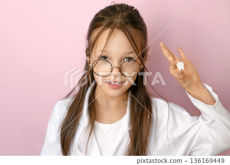 Smiling child in medical coat holding first baby tooth on pink background, concept of National Children Dental Health awareness, milk teeth loss, pediatric dentistry, oral hygiene education and preven Smiling child in medical coat holding first baby tooth on pink background, concept of National Children Dental Health awareness, milk teeth loss, pediatric dentistry, oral hygiene education and preven 136169449
