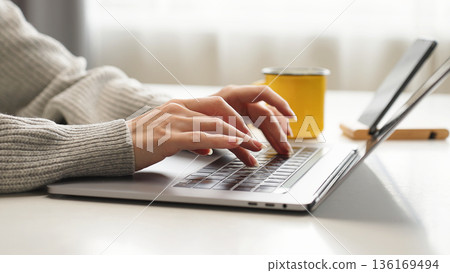 Close-up of woman's hands typing on a laptop at home, working on email, business report, data entry, research, and remote communication for company tasks. Close-up of woman's hands typing on a laptop at home, working on email, business report, data entry, research, and remote communication for company tasks. 136169494