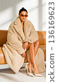A woman sitting on a couch wearing a beige teddy bear coat 136169623