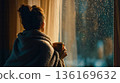 A woman wrapped in a blanket looking out a window with a cup of coffee 136169632