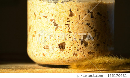 Close-up of fermenting rye sourdough with a bubbly texture, showcasing wild yeast in home baking. Starter in a glass jar with wheat stalk, demonstrating active yeast. Healthy fermented food commercial 136170681