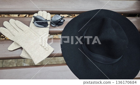 Autumn fashion accessories arranged on a wooden park bench with black hat sunglasses and beige gloves. Concept of seasonal style minimalism and urban elegance 136170684