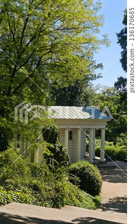 Classical pavilion with white columns surrounded by lush green trees in a sunny park under blue sky. Concept of harmony between architecture nature and peaceful outdoor recreation 136170685