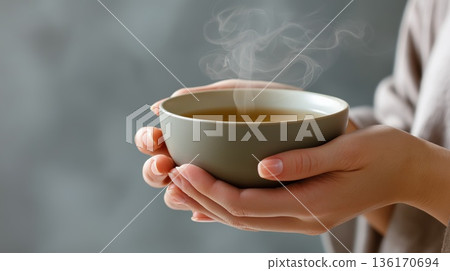 Warm cup of tea gently held in human hands with visible steam against a soft neutral background. Concept of comfort relaxation mindfulness and cozy everyday ritual 136170694