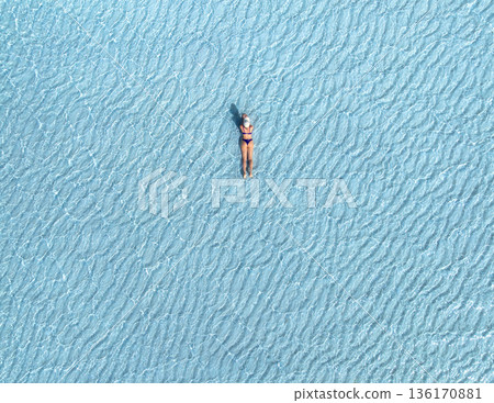 Aerial view of woman in bikini and white hat lying in blue sea 136170881
