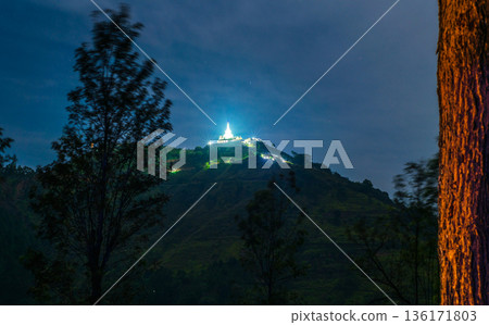 Illuminated hilltop Buddhist temple glows above forested montane slopes in Nuwara Eliya, Sri Lanka. Long exposure night photograph. Illuminated hilltop Buddhist temple glows above forested montane slopes in Nuwara Eliya, Sri Lanka. Long exposure night photograph. 136171803