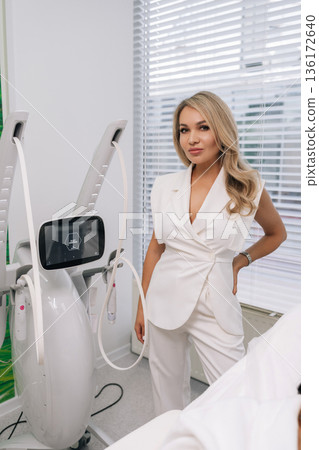 Professional female cosmetologist working in contemporary medical aesthetic clinic, standing confidently by advanced SMAS lifting equipment for facial rejuvenation and body contouring procedures. 136172640