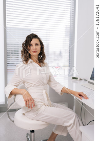 Confident female doctor in bright modern clinic office looking at camera sitting at desk using computer, professional and approachable portrait conveying medical expertise and reliability. 136172641