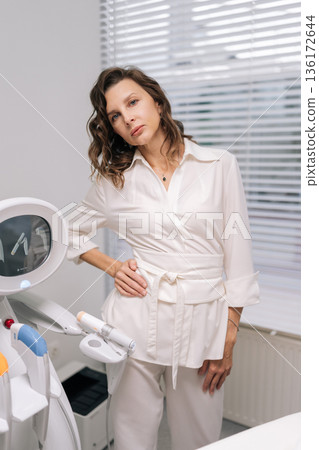 Professional cosmetologist in white uniform standing next to advanced SMAS lifting machine in clean beauty clinic, preparing for procedure offering skincare and anti-aging treatments. Professional cosmetologist in white uniform standing next to advanced SMAS lifting machine in clean beauty clinic, preparing for procedure offering skincare and anti-aging treatments. 136172644