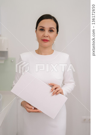 Cosmetologist in clean clinic holding blank white box for branding or product display, conveying professional medical aesthetics, skincare expertise and calm confidence. Concept of beauty treatment. Cosmetologist in clean clinic holding blank white box for branding or product display, conveying professional medical aesthetics, skincare expertise and calm confidence. Concept of beauty treatment. 136172650
