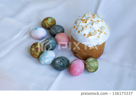 Orthodox Easter kulich decorated with cashew nuts and icing on a white tablecloth with painted eggs 136173337