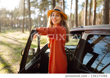 Young woman enjoying a sunny day by her car in forest setting during the afternoon. Summer trip Young woman enjoying a sunny day by her car in forest setting during the afternoon. Summer trip 136173366
