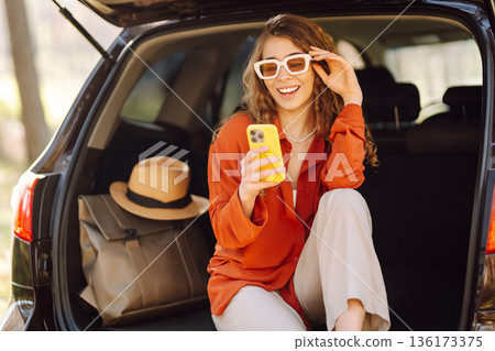 Beautiful woman sits in the trunk of a car outdoor and uses a smartphone. Active life. Beautiful woman sits in the trunk of a car outdoor and uses a smartphone. Active life. 136173375
