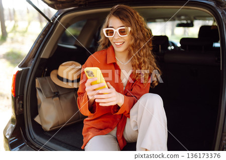 Beautiful woman sits in the trunk of a car outdoor and uses a smartphone. Active life. Beautiful woman sits in the trunk of a car outdoor and uses a smartphone. Active life. 136173376