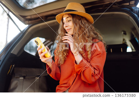 Beautiful woman sits in the trunk of a car outdoor and uses a smartphone. Active life. 136173380