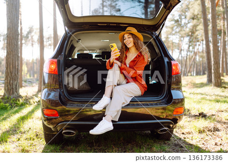 Beautiful woman sits in the trunk of a car outdoor and uses a smartphone. Active life. Beautiful woman sits in the trunk of a car outdoor and uses a smartphone. Active life. 136173386