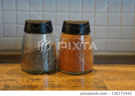 front view put chili powder in a glass cup With a black cover and ground pepper in a glass cup With a black cover, placed on a wooden floor, white ceramic tile wall background, copy space 136173442