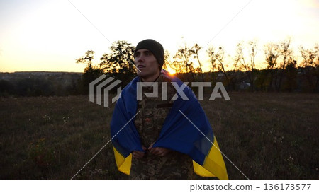 Soldier of ukrainian army going through meadow with Ukraine flag as symbol of victory against russian aggression. Young male military in uniform walking with blue-yellow banner at countryside Soldier of ukrainian army going through meadow with Ukraine flag as symbol of victory against russian aggression. Young male military in uniform walking with blue-yellow banner at countryside 136173577