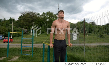 Young sportsman doing push ups on parallel bars at nature. Strong sporty man exercising at sports ground. Male shirtless athlete training hard outdoor. Concept of active sportive lifestyle. Slow mo Young sportsman doing push ups on parallel bars at nature. Strong sporty man exercising at sports ground. Male shirtless athlete training hard outdoor. Concept of active sportive lifestyle. Slow mo 136173585