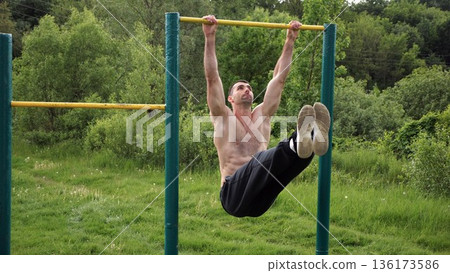 Athletic man trains abdominal muscles raising legs on horizontal bar at sports ground. Young sportsman exercising on pull-up bar outdoor. Sporty athlete working out at nature. Healthy lifestyle 136173586