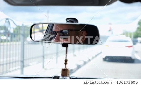 Eyes in sunglasses of male driver reflecting in rearview mirror while riding motor auto. Young guy looking concentrated to urban road during operating vehicle. Confident man driving modern car at city 136173609