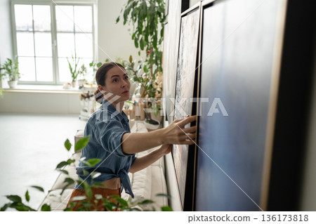 Thoughtful talented woman painter carefully adjusting hanging painting on wall at studio gallery. Thoughtful talented woman painter carefully adjusting hanging painting on wall at studio gallery. 136173818
